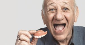 An elderly man smiling while holding his dentures in his hand